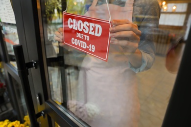 Woman putting red sign with words "Closed Due To Covid-19" onto glass door, closeup