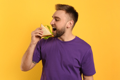 Young man eating tasty sandwich on yellow background