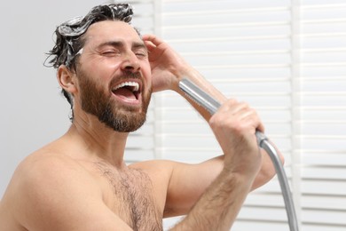 Happy man with showerhead singing and washing his hair with shampoo indoors