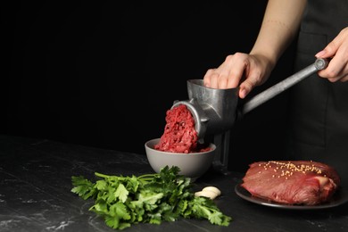 Woman making beef mince with manual meat grinder at dark textured table against black background, closeup. Space for text