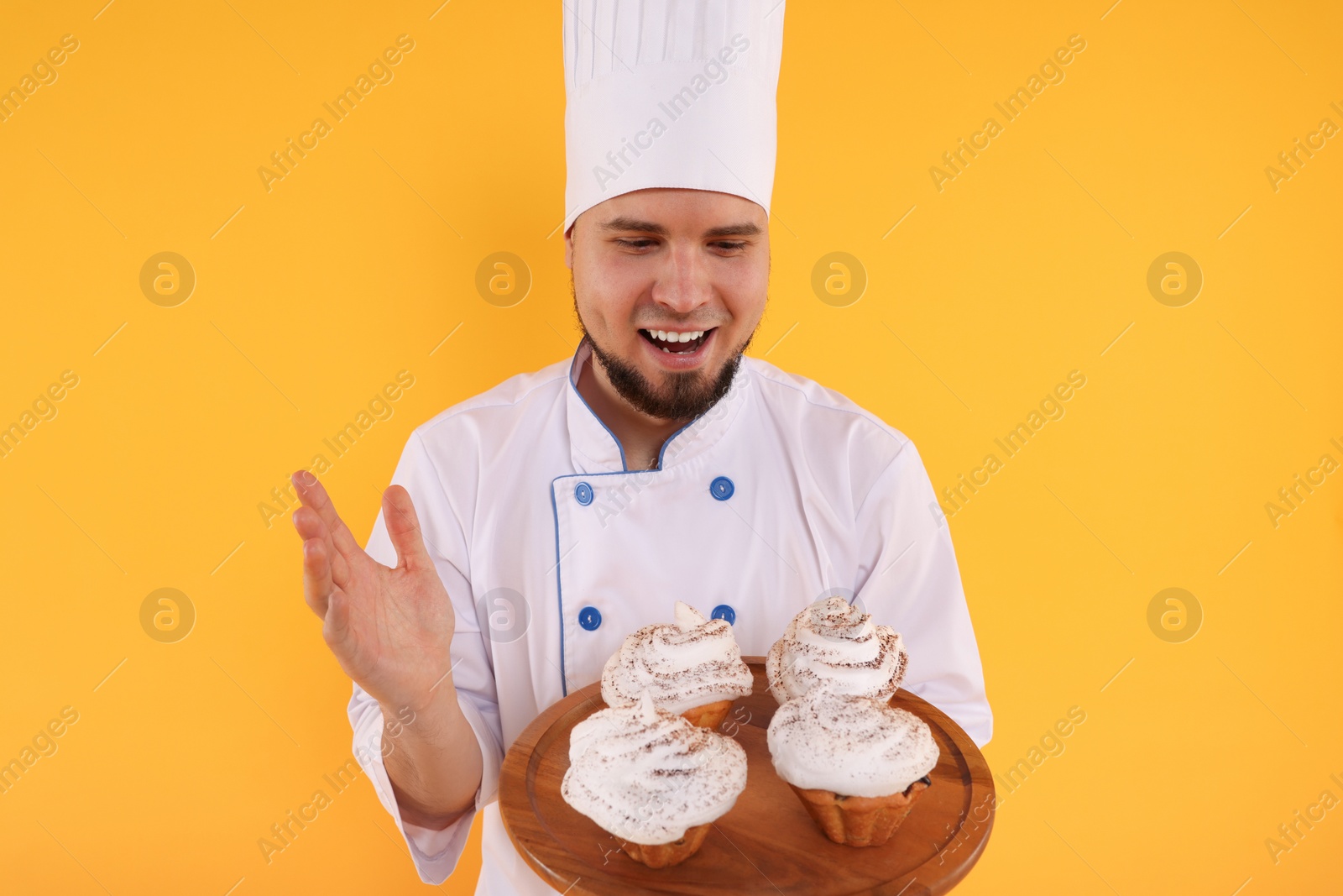 Photo of Happy professional confectioner in uniform holding delicious cupcakes on yellow background