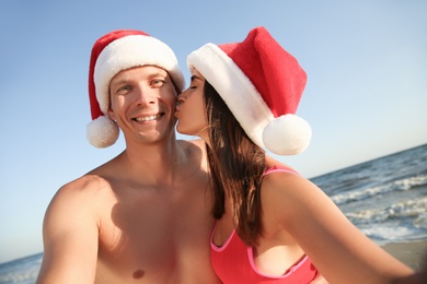 Photo of Happy couple with Santa hats taking selfie together on beach. Christmas vacation