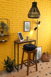 Photo of High wooden table with tablet and stool as stand up workplace near yellow brick wall. Stylish interior