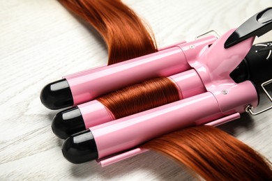 Curling iron with red hair lock on white wooden table, closeup