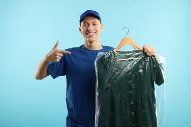 Dry-cleaning delivery. Happy courier holding dress in plastic bag on light blue background