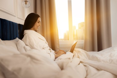 Image of Happy young woman working with laptop on bed in hotel room