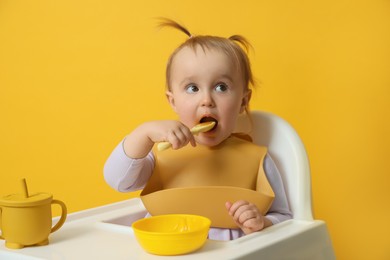 Cute little baby wearing bib while eating on yellow background