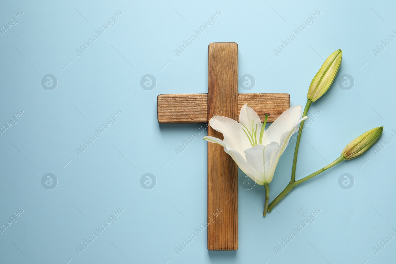 Photo of Wooden cross and lily flowers on light blue background, top view with space for text. Easter attributes