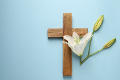 Photo of Wooden cross and lily flowers on light blue background, top view with space for text. Easter attributes