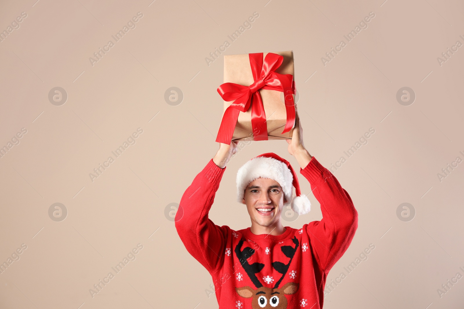 Photo of Happy man in Christmas sweater and Santa hat holding gift box on beige background