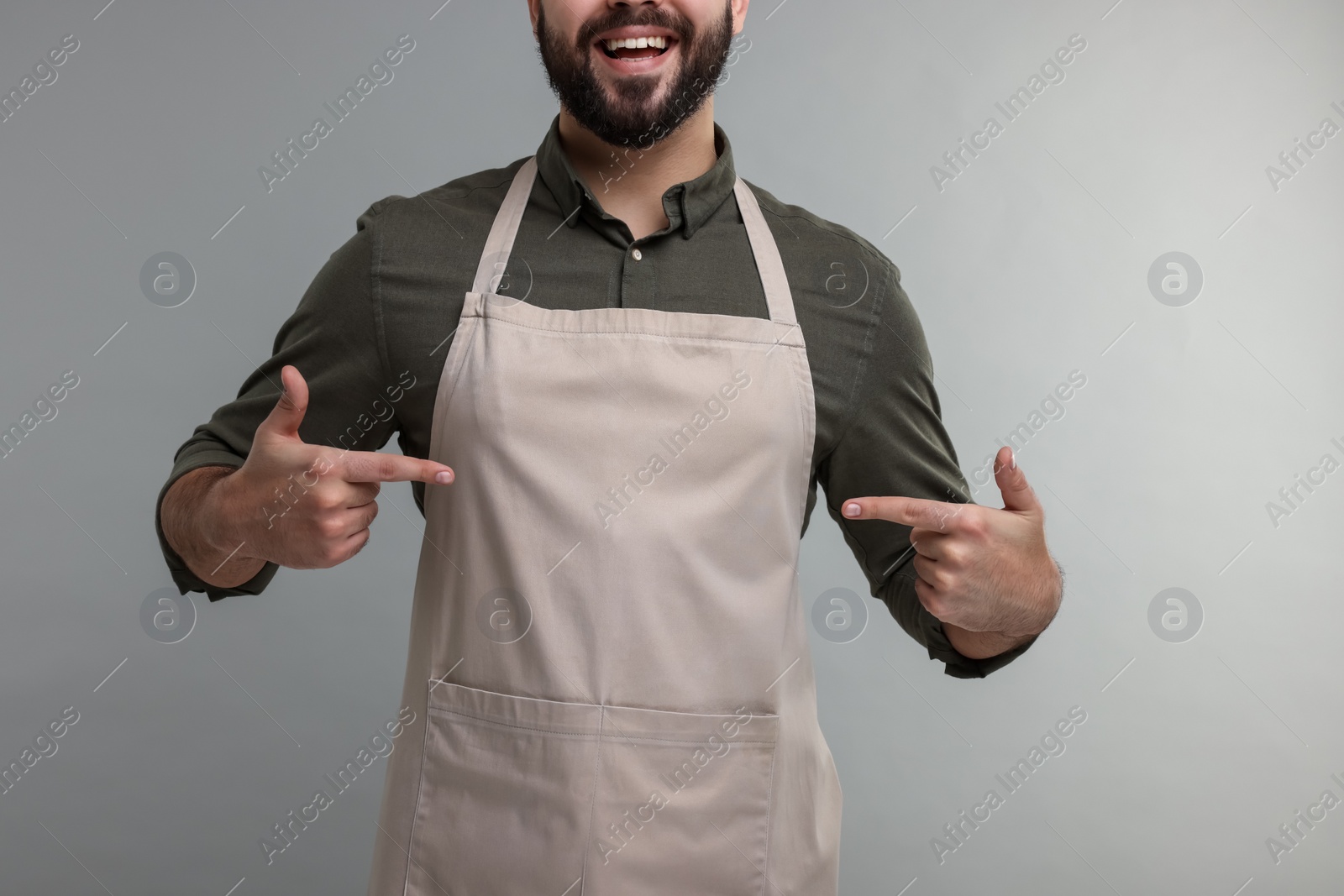 Photo of Happy man pointing at kitchen apron on grey background, closeup. Mockup for design
