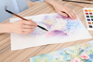 Photo of Woman painting flowers with watercolor at wooden table, closeup. Creative artwork