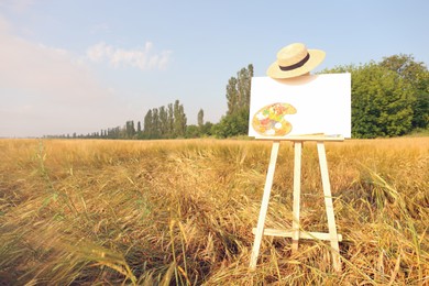 Photo of Wooden easel with blank canvas, painting equipment and hat in field. Space for text