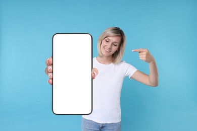 Happy woman pointing at mobile phone with blank screen on light blue background. Mockup for design