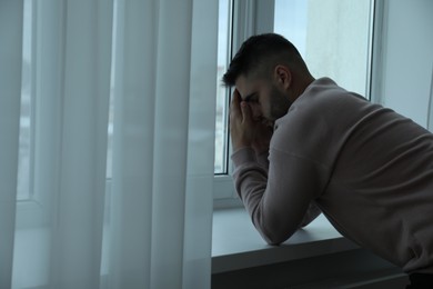 Photo of Sad man near window at home. Space for text