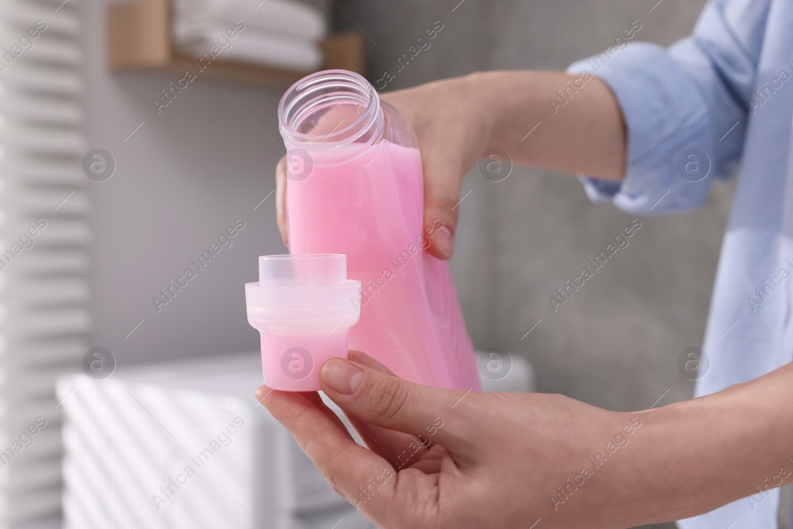 Photo of Woman pouring fabric softener from bottle into cap for washing clothes indoors, closeup