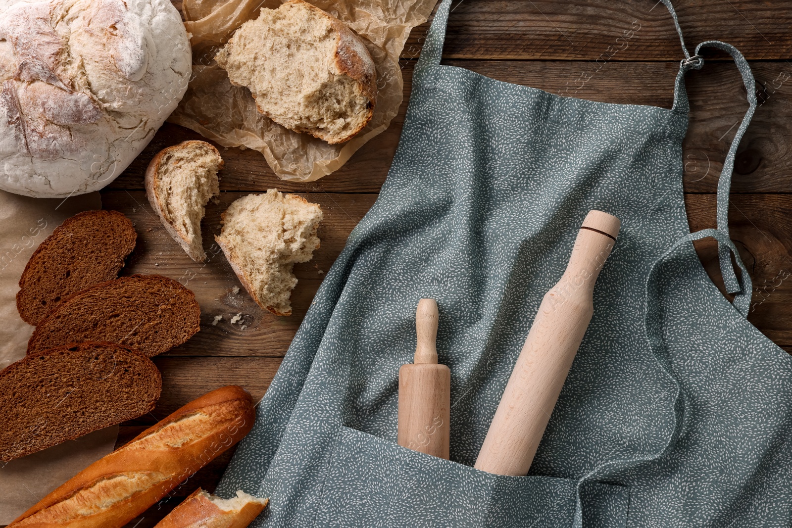 Photo of Clean kitchen apron with rolling pins and different types of bread on wooden table, flat lay