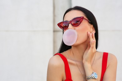 Photo of Beautiful woman in stylish sunglasses blowing gum near wall outdoors, space for text