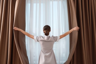 Chambermaid opening window curtains in hotel room, back view