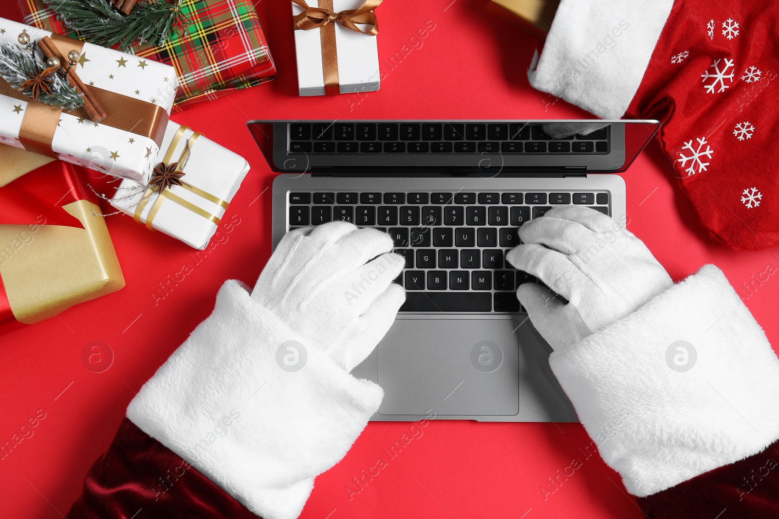 Photo of Santa Claus using laptop, closeup. Gift boxes and Christmas decor on red background, top view