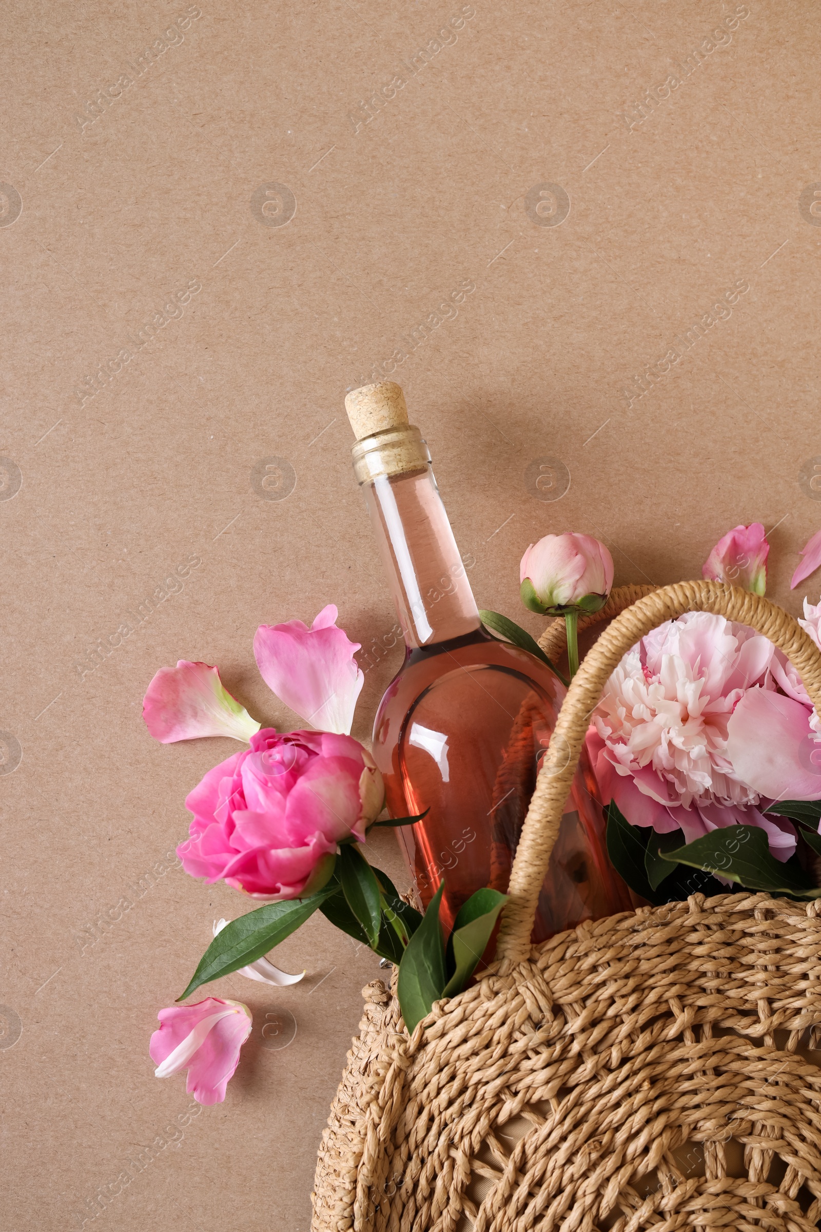 Photo of Wicker bag with bottle of rose wine and beautiful pink peonies on brown background, top view. Space for text