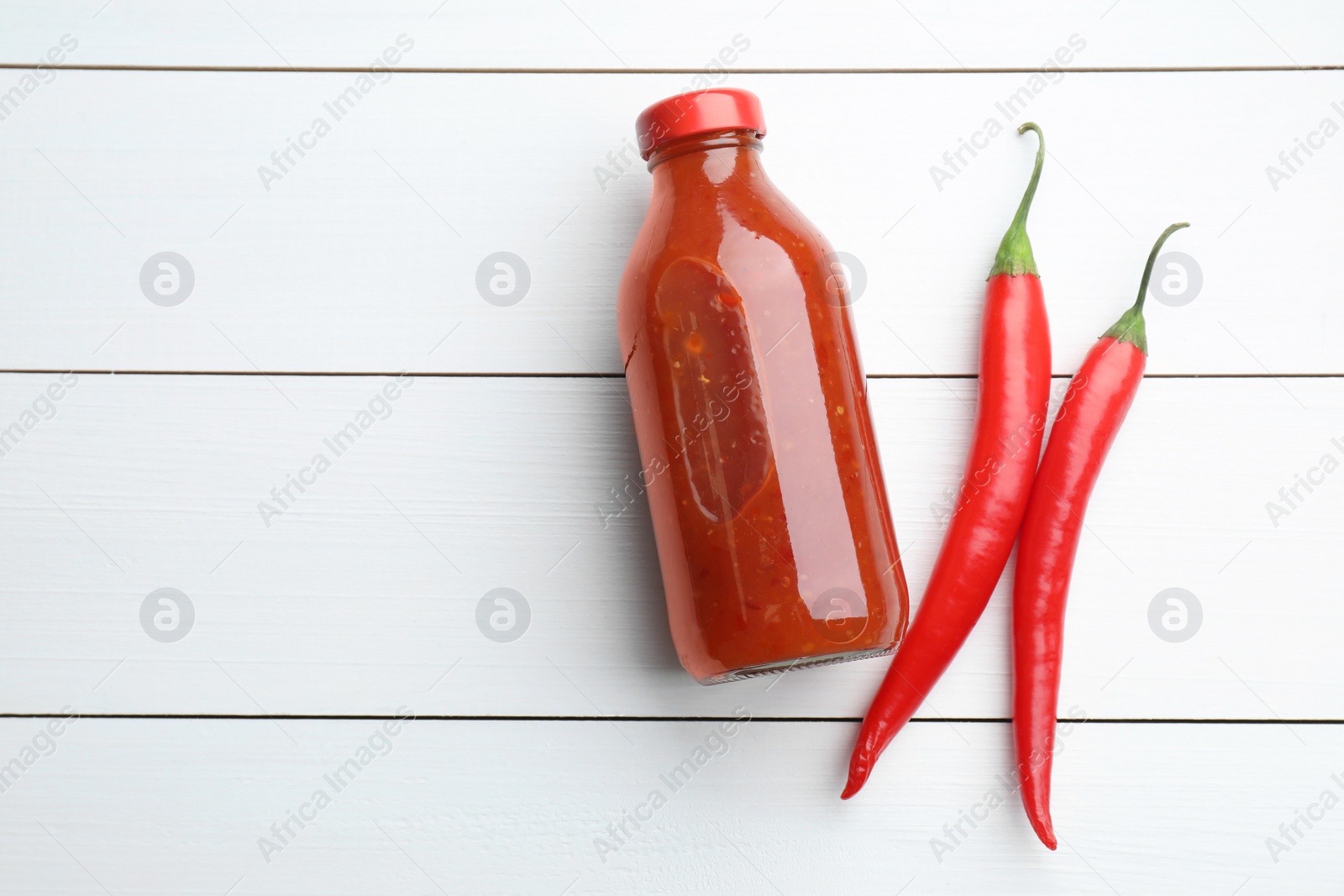 Photo of Spicy chili sauce in bottle and peppers on white wooden table, flat lay. Space for text