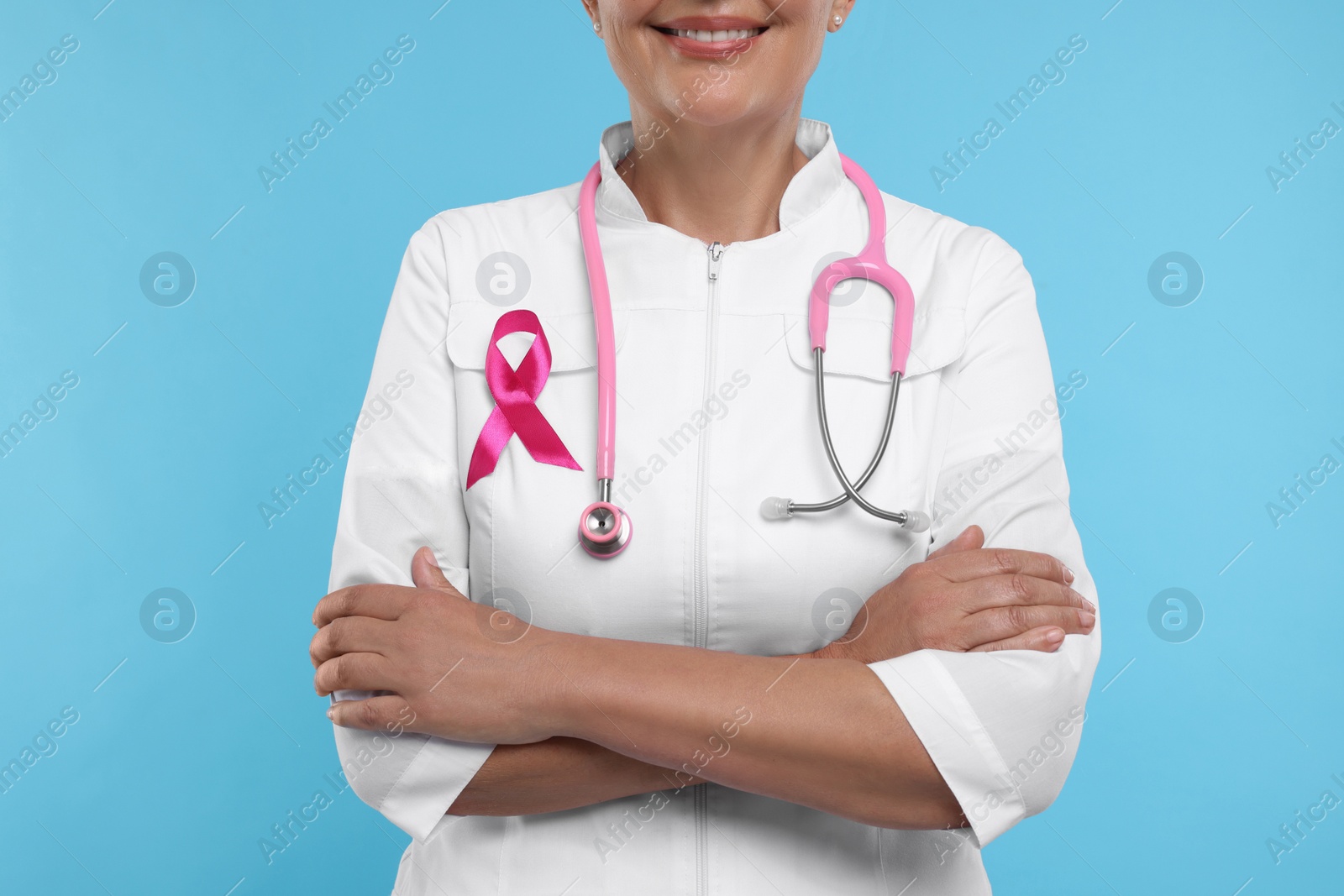 Photo of Doctor with pink ribbon and stethoscope on light blue background, closeup. Breast cancer awareness