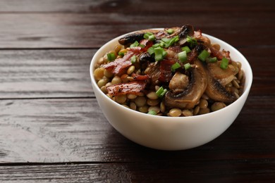 Photo of Delicious lentils with mushrooms, bacon and green onion in bowl on wooden table, closeup. Space for text