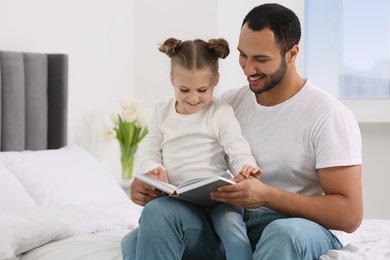 Little girl with her father reading book together on bed at home. International family