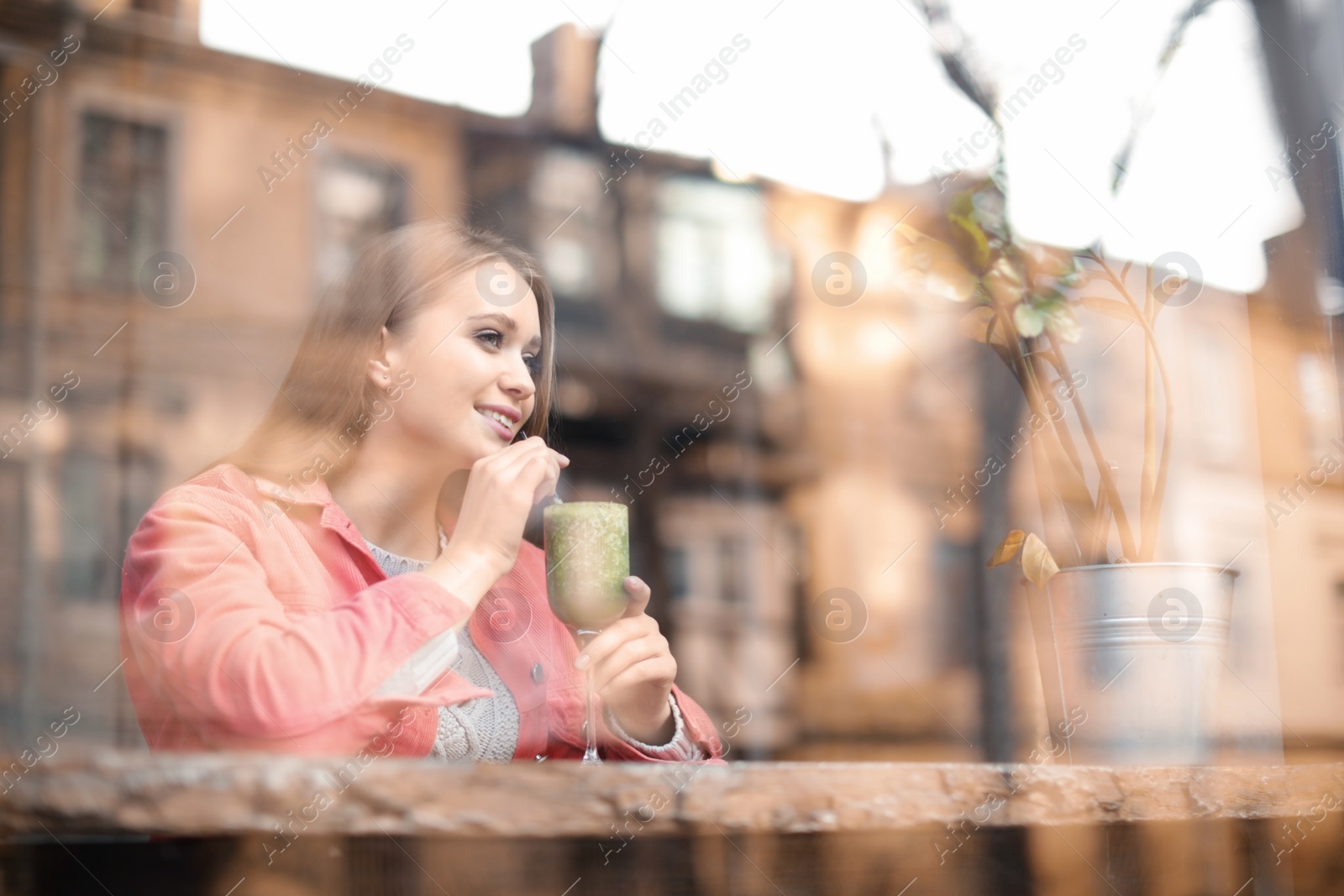 Photo of Pretty young woman with cocktail at table in cafe, view from outdoors through window