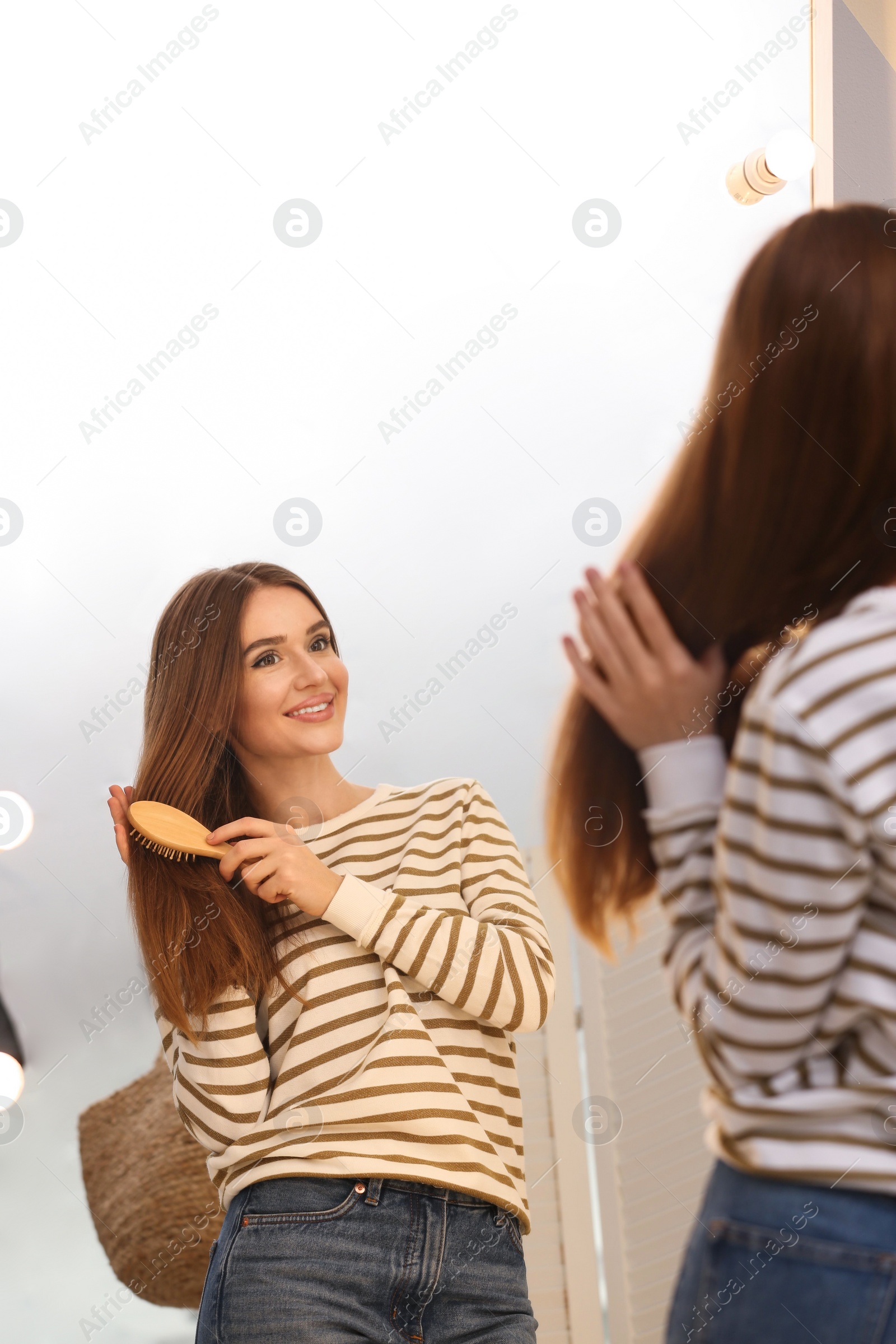 Photo of Beautiful young woman with hair brush looking at herself in large mirror indoors