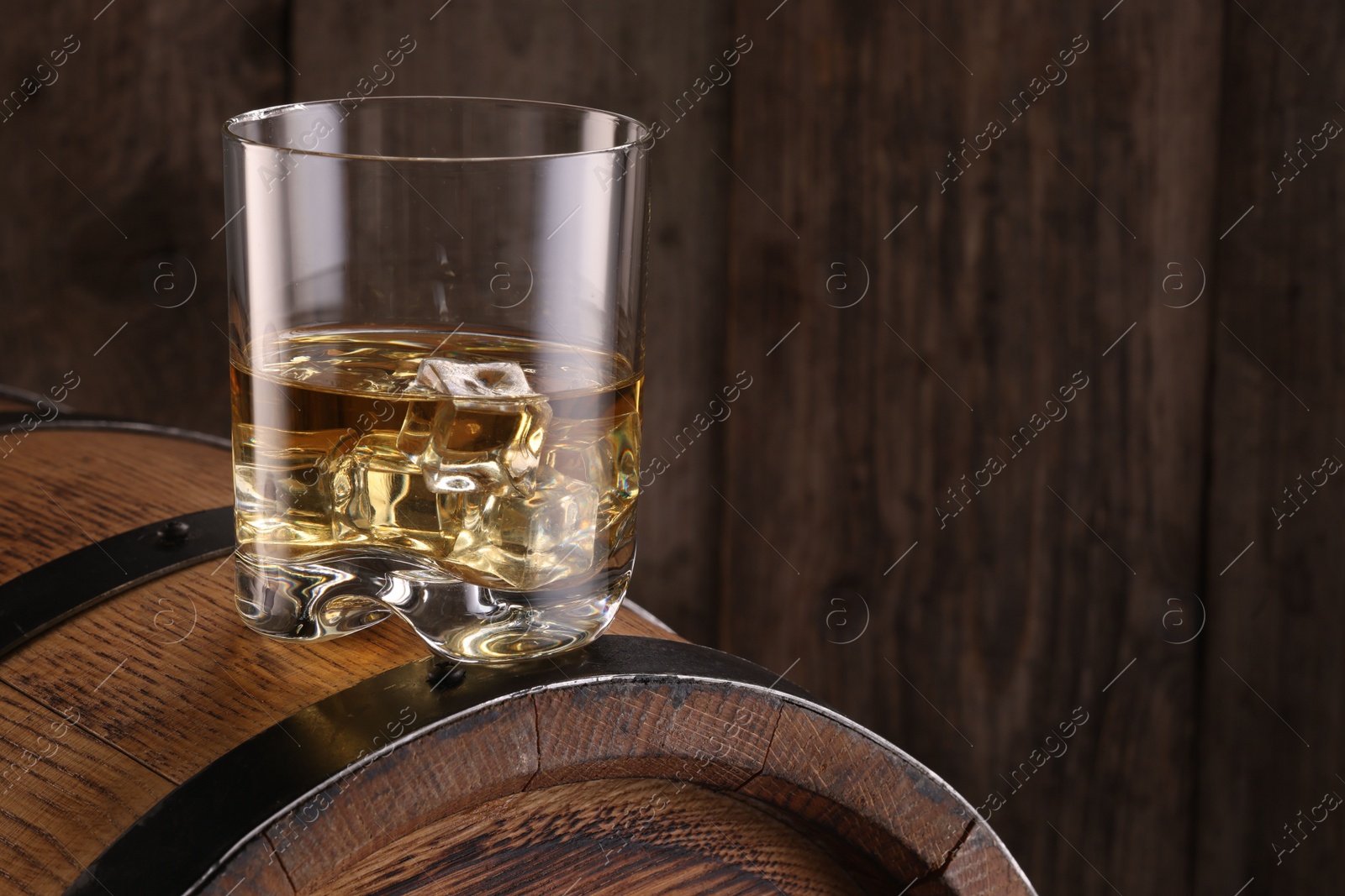 Photo of Whiskey with ice cubes in glass on barrel against wooden background, space for text