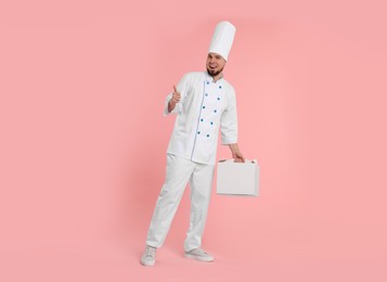 Photo of Happy professional confectioner in uniform holding cake box and showing thumbs up on pink background