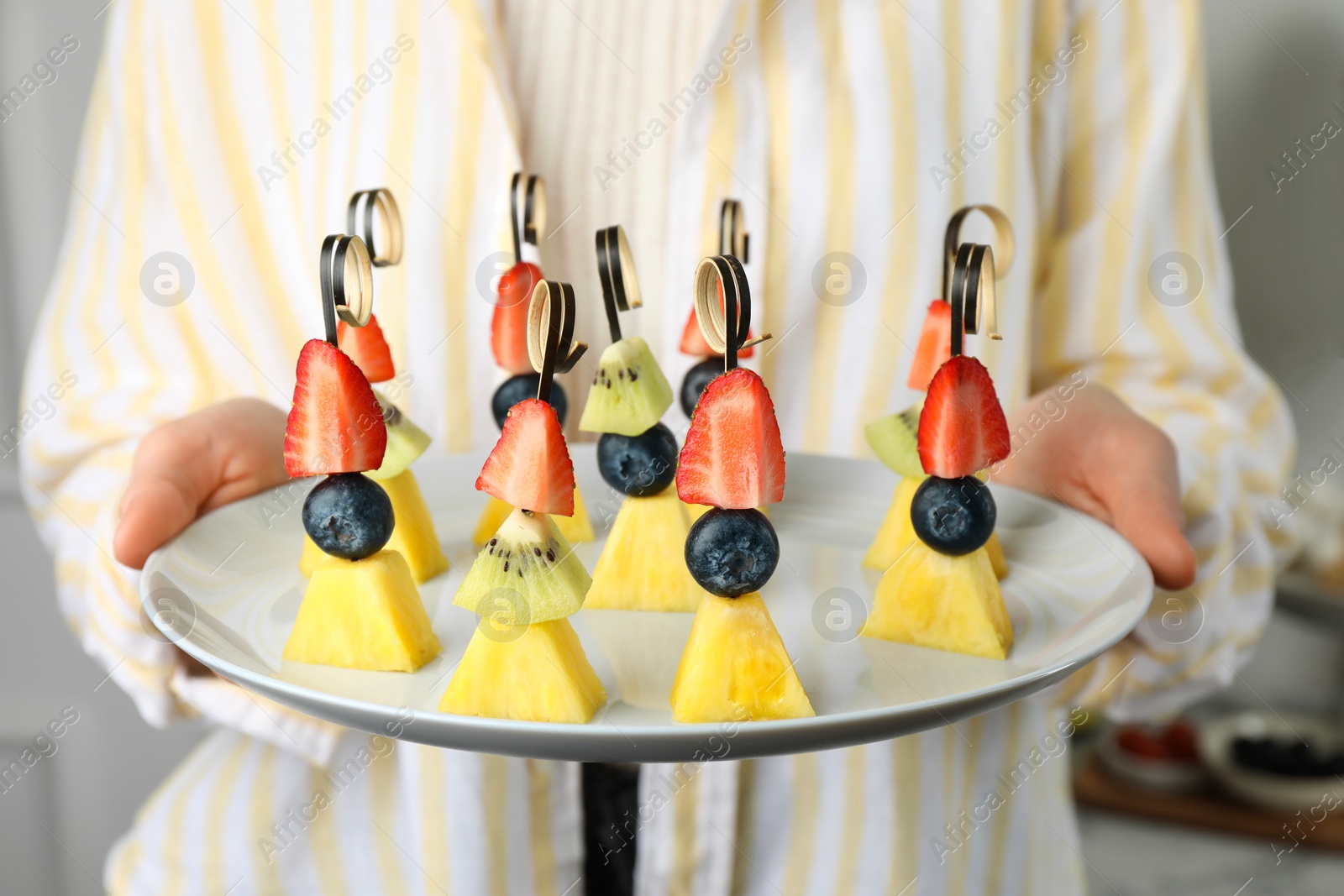 Photo of Woman holding plate of tasty canapes with pineapple, kiwi and berries indoors, closeup