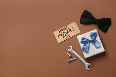 Photo of Card with phrase HAPPY FATHER'S DAY, tools, bow tie and gift box on brown background, flat lay. Space for text