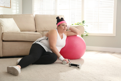 Photo of Lazy overweight woman with sport equipment on floor at home