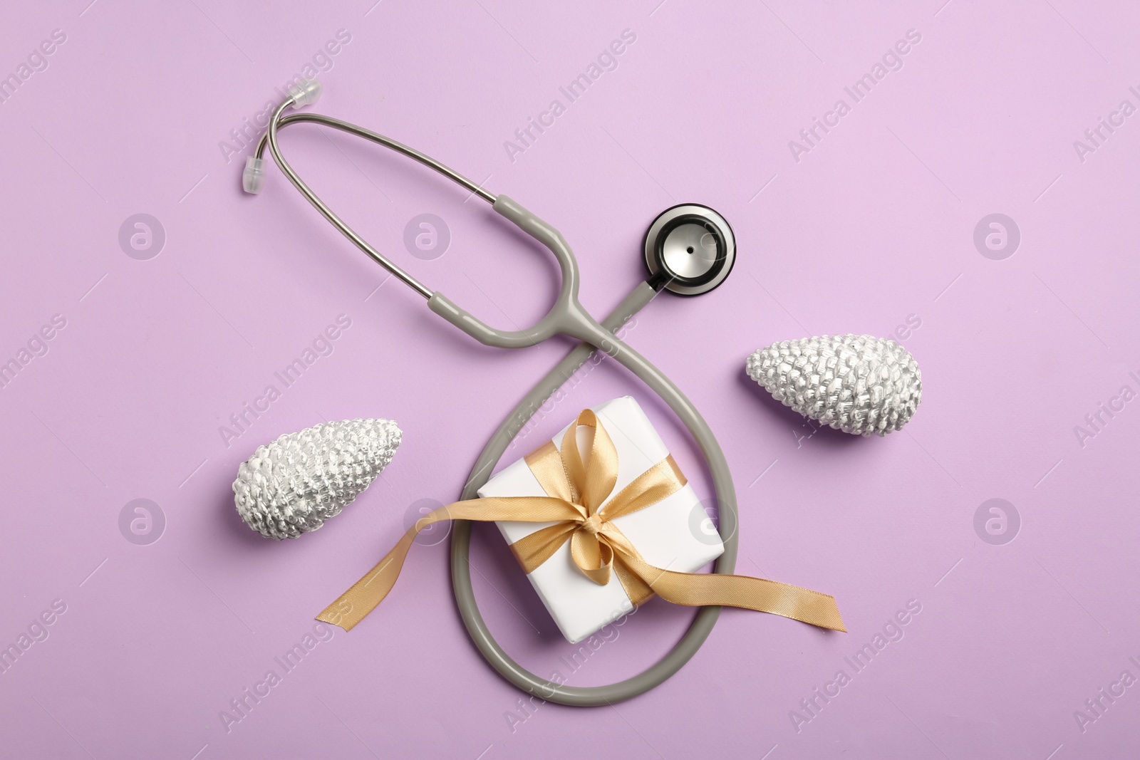 Photo of Greeting card for doctor with stethoscope, gift box and Christmas decor on purple background, flat lay