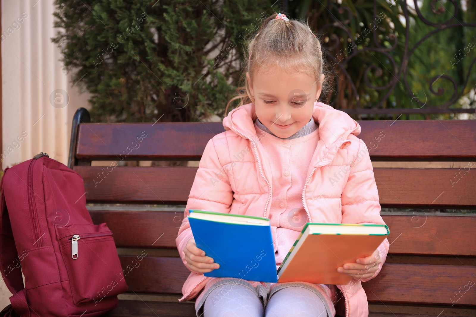 Photo of Cute little girl with backpack and books on bench outdoors