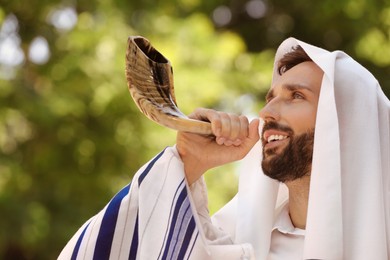 Photo of Jewish man in tallit blowing shofar outdoors. Rosh Hashanah celebration