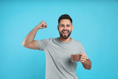 Photo of Young man with glass cup of chocolate milk showing his strength on light blue background
