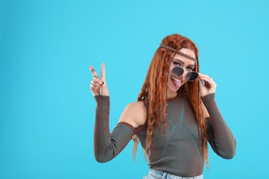 Stylish young hippie woman in sunglasses showing V-sign on light blue background, space for text