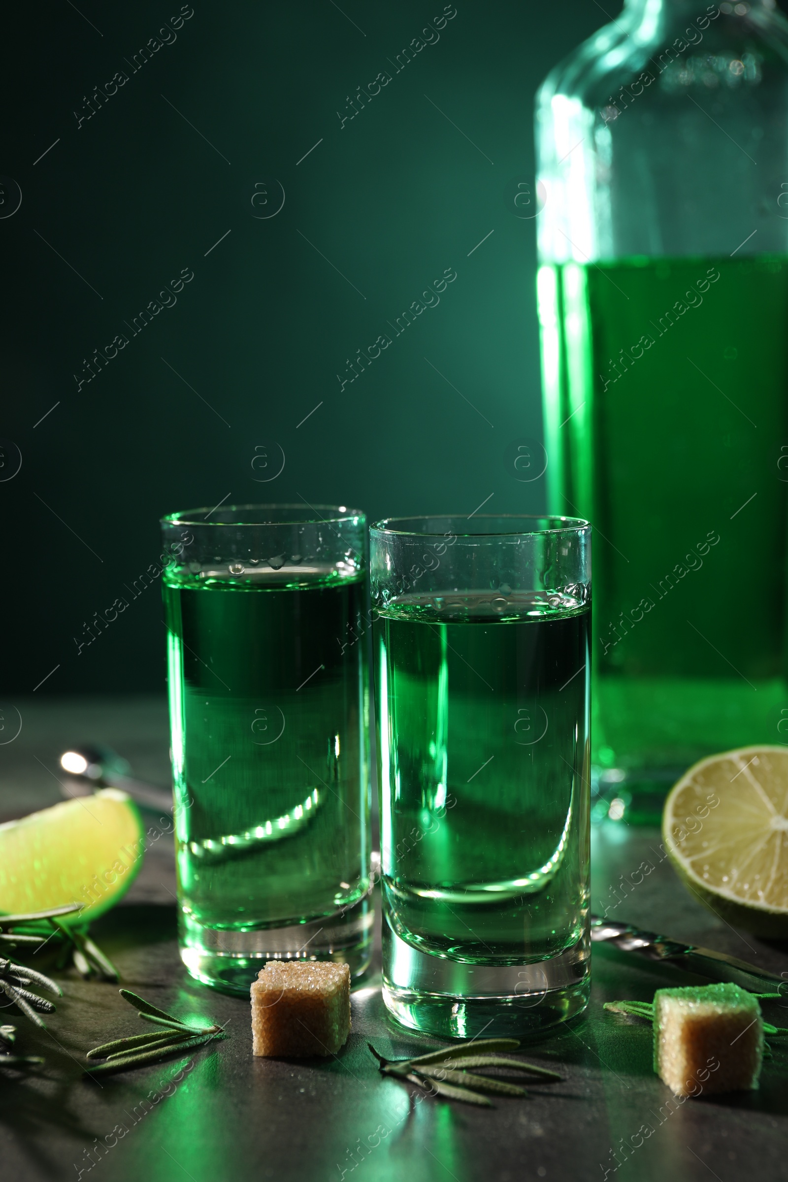 Photo of Absinthe in shot glasses, brown sugar cubes and rosemary on gray table, closeup. Alcoholic drink