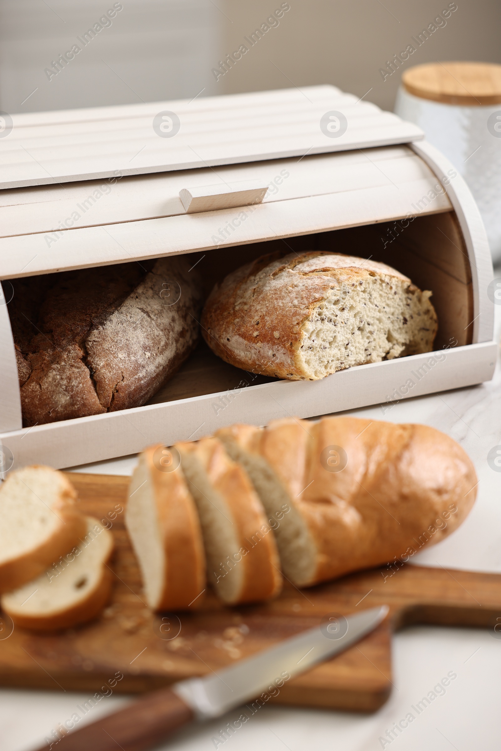 Photo of Wooden bread basket with freshly baked loaves and knife on white marble table in kitchen