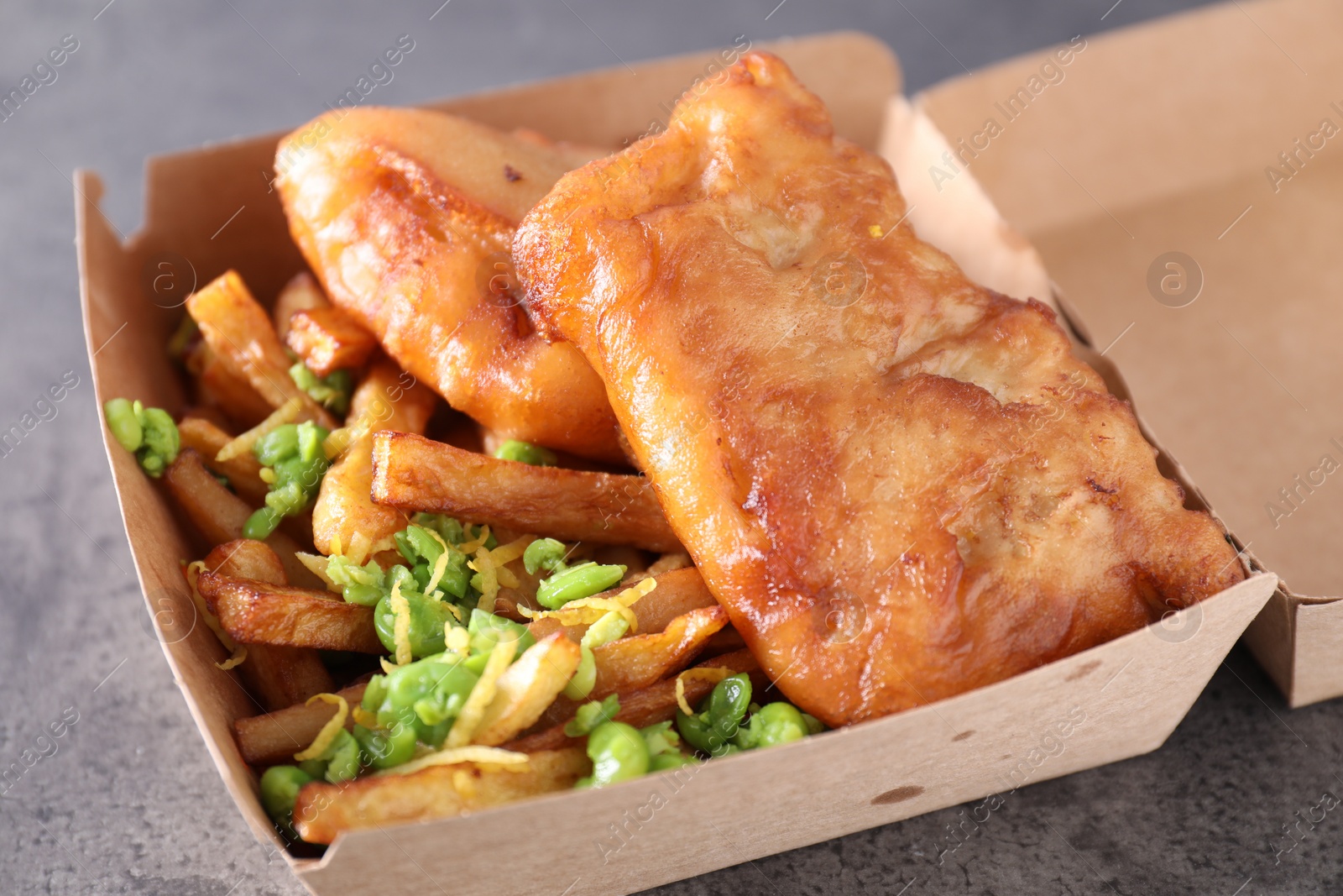 Photo of Tasty fish, chips and peas in paper box on grey table, closeup