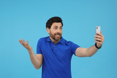 Puzzled man taking selfie with smartphone on light blue background