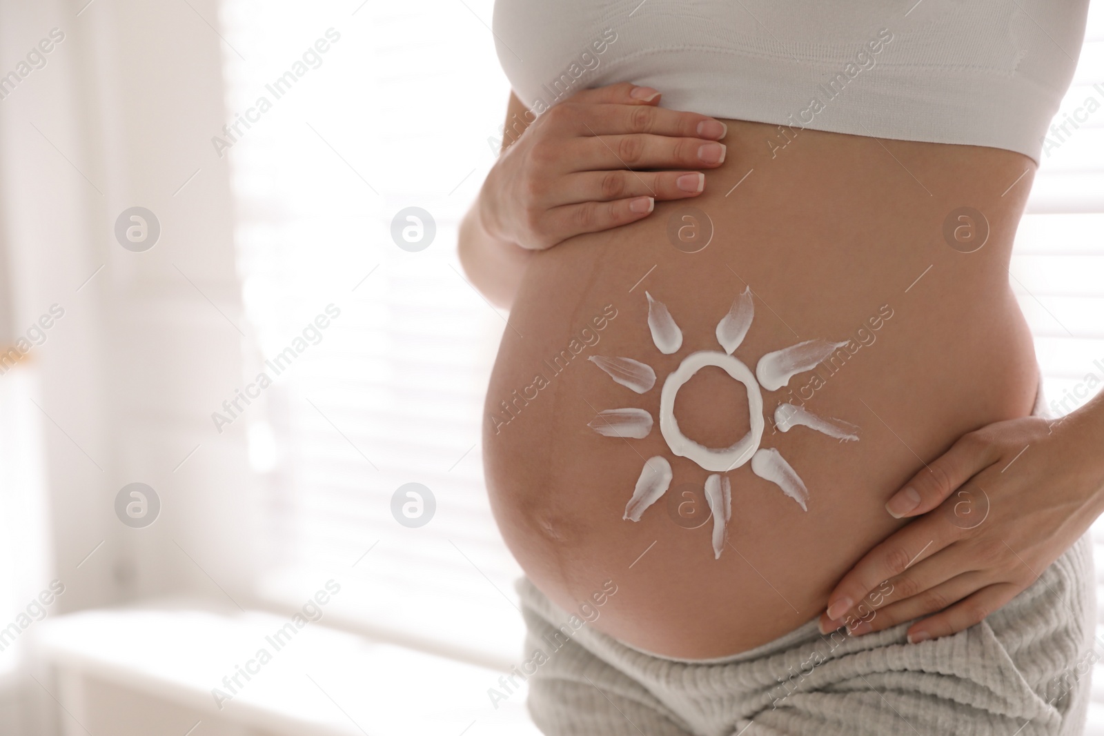 Photo of Young pregnant woman with sun protection cream on belly indoors, closeup. Space for text