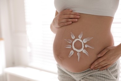 Photo of Young pregnant woman with sun protection cream on belly indoors, closeup. Space for text