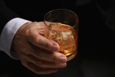 Photo of Man holding glass of whiskey with ice cubes on dark background, closeup
