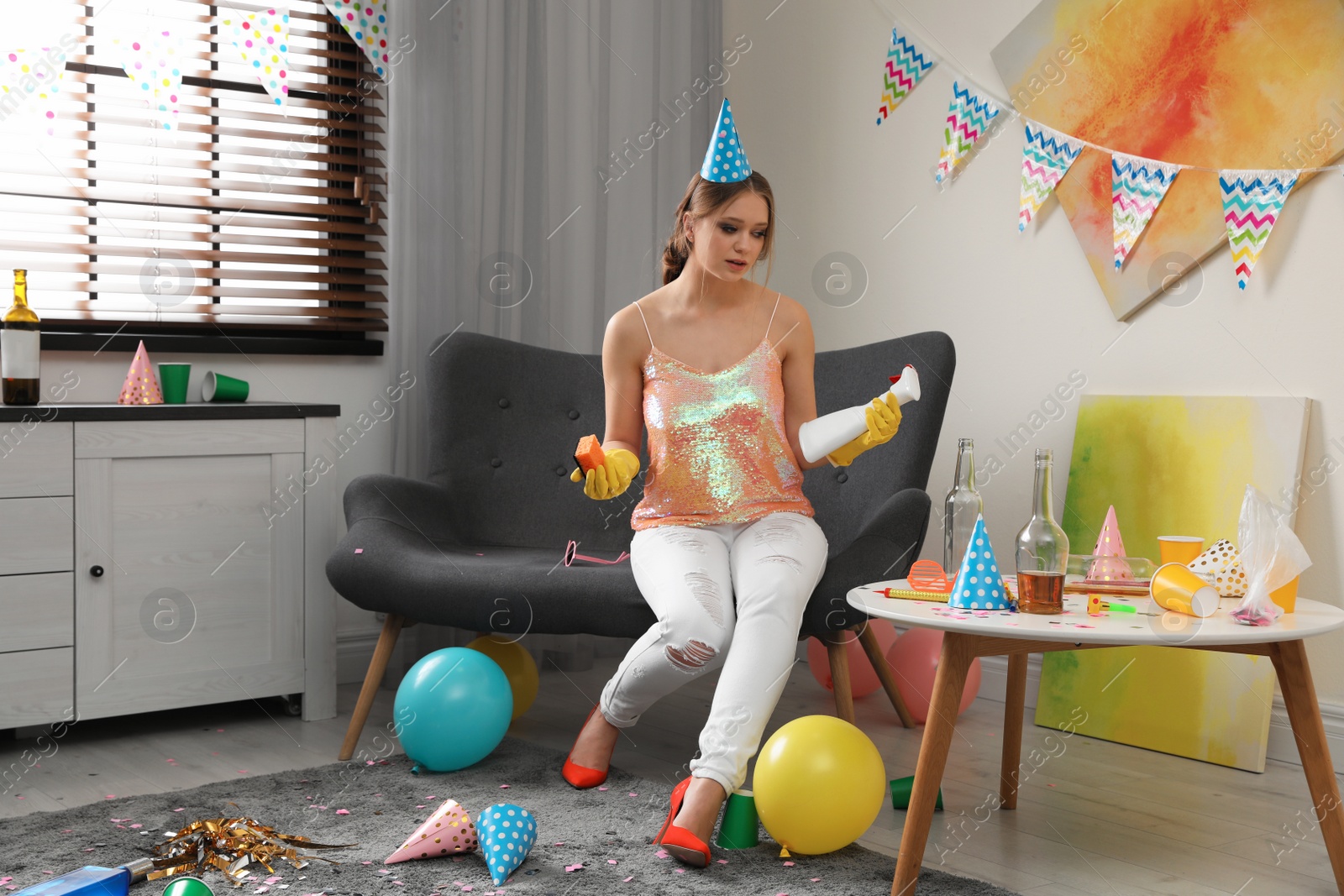 Photo of Tired young woman with bottle of detergent and sponge in messy room after party