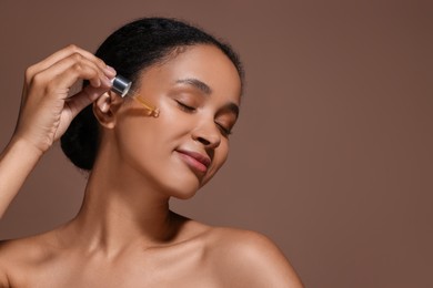 Beautiful woman applying serum onto her face on brown background. Space for text